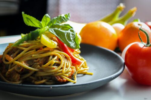 Pasta with vegetable dish on gray plate beside tomato fruit on white table