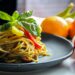 Pasta with vegetable dish on gray plate beside tomato fruit on white table